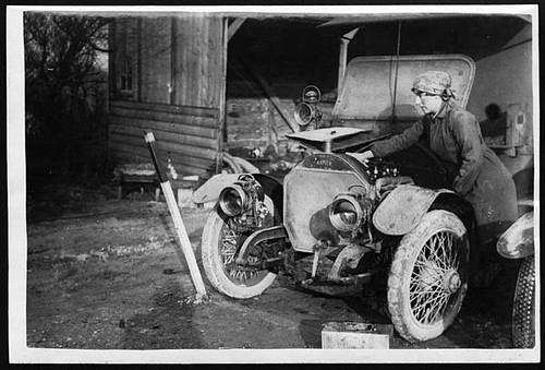 A young woman, a member of the First Aid Nursing Yeomanry, oils her car engine, ina black and white image from early 20th century