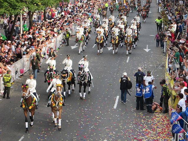 Military on horseback, as a woman who may have been suffering from exhaustion is carried away on a stretcher (right side).