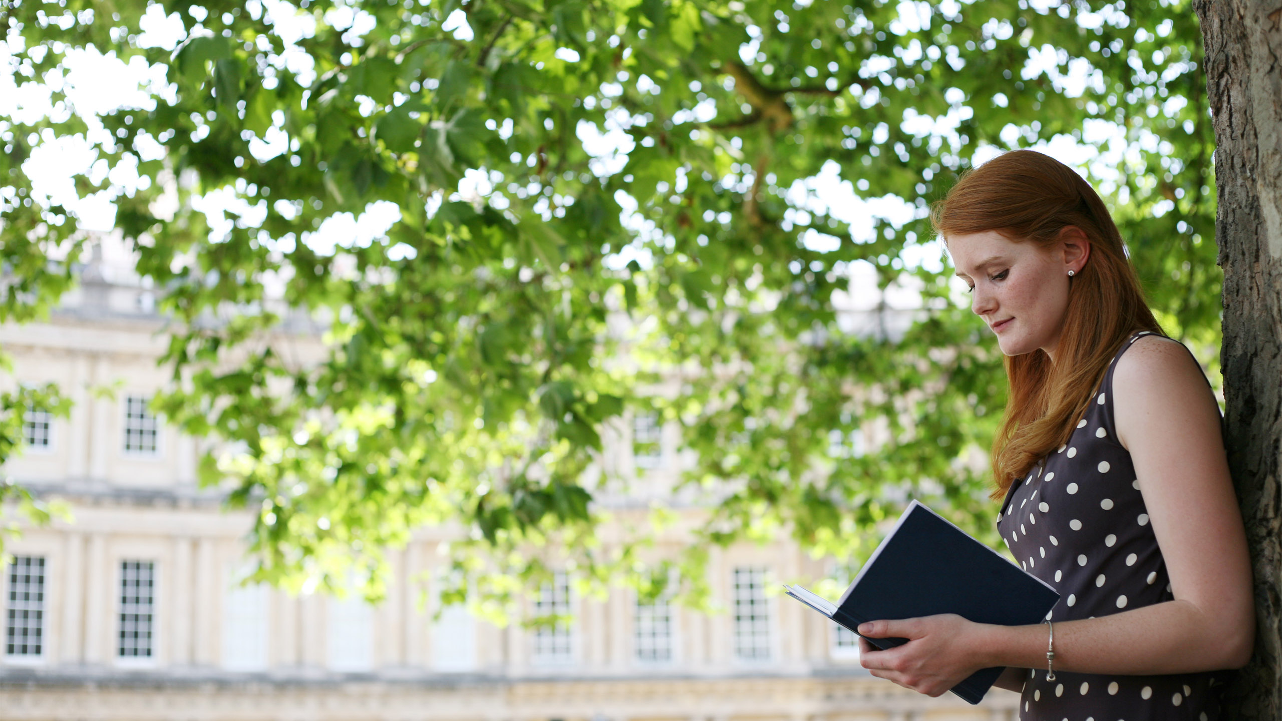 在全球官网电子游戏广场，一名学生靠在一棵树上看书