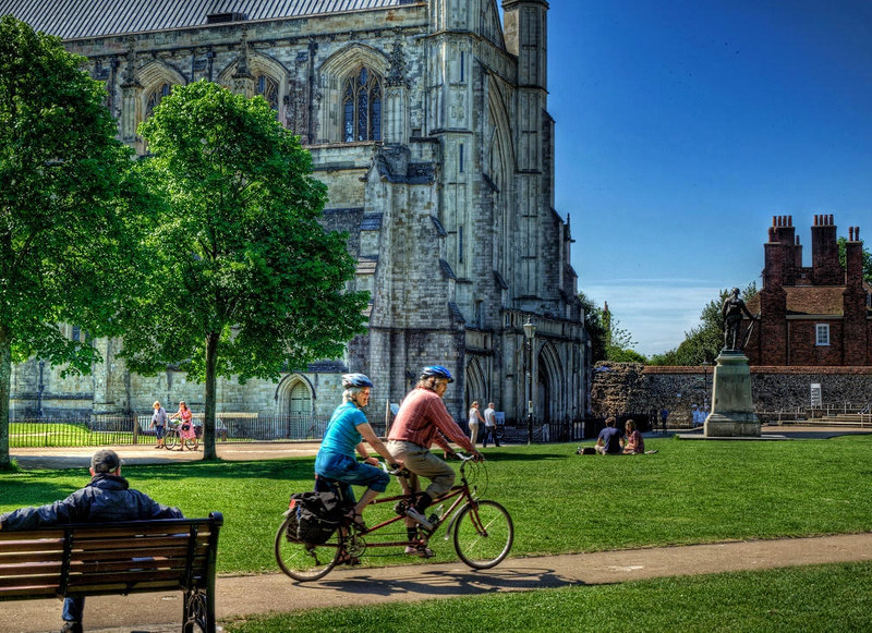 A couple cycle past Winchester Cathedral on a bicycle made for two. Credit Neil Howard, flickr