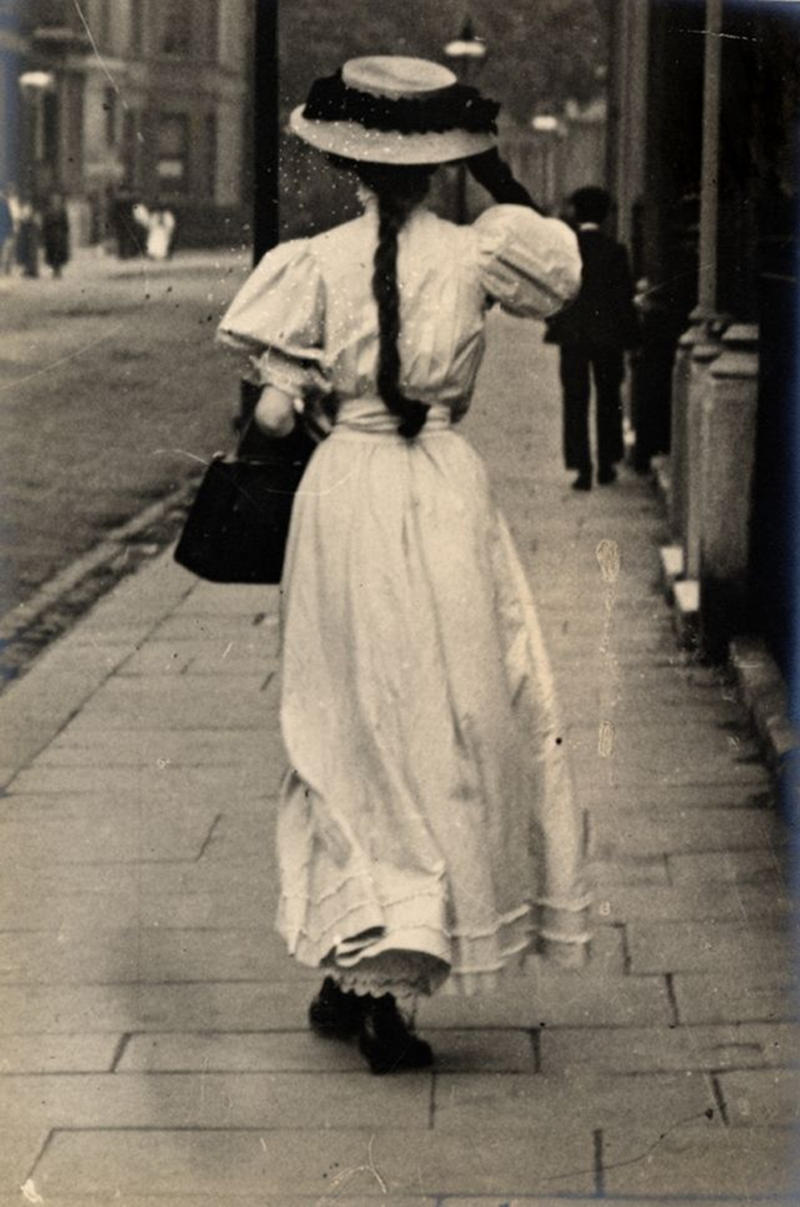 A woman in a formal white dress with black handbag walks along the street in Kensington on June 15, 1908