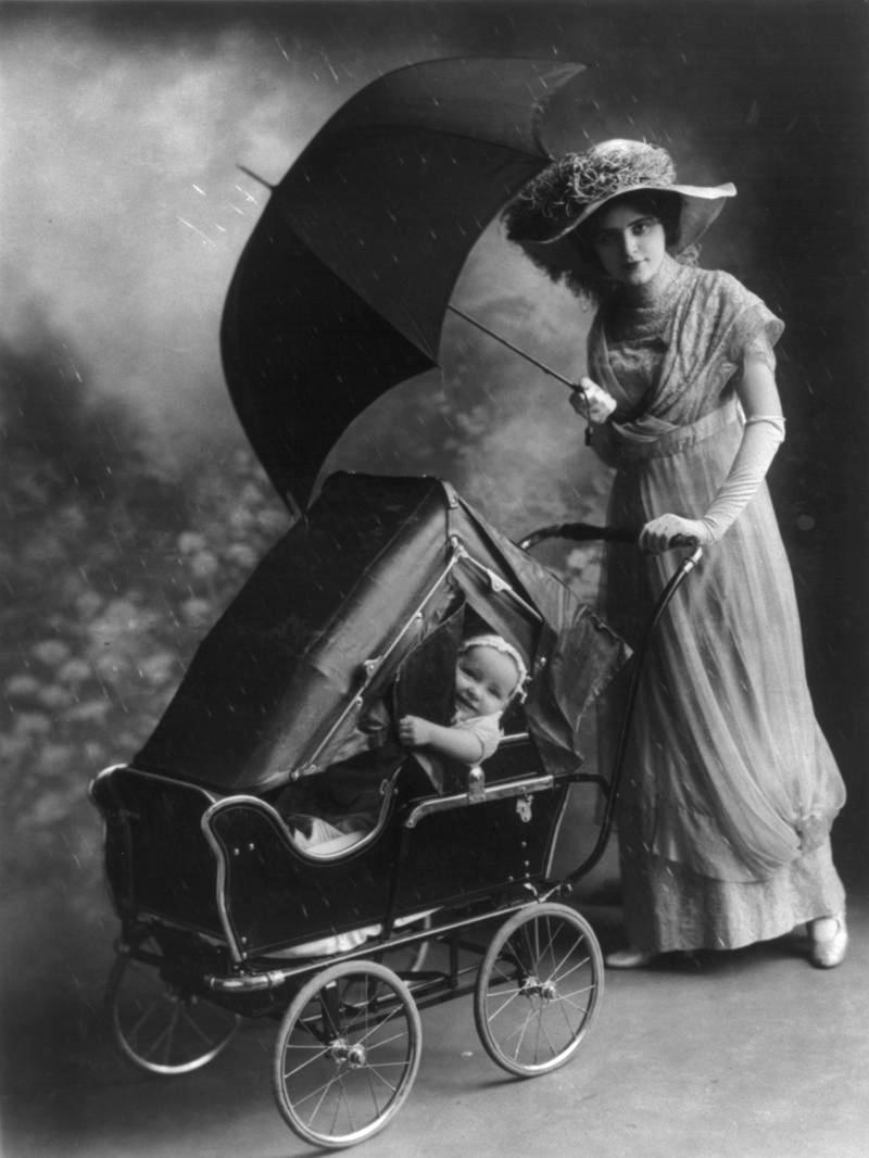 Woman, holding umbrella, pushing baby in carriage equipped with rain cover