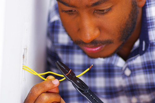 Man wearing white and blue shirt working on electrical wall socket wires using screwdriver, electrician concept