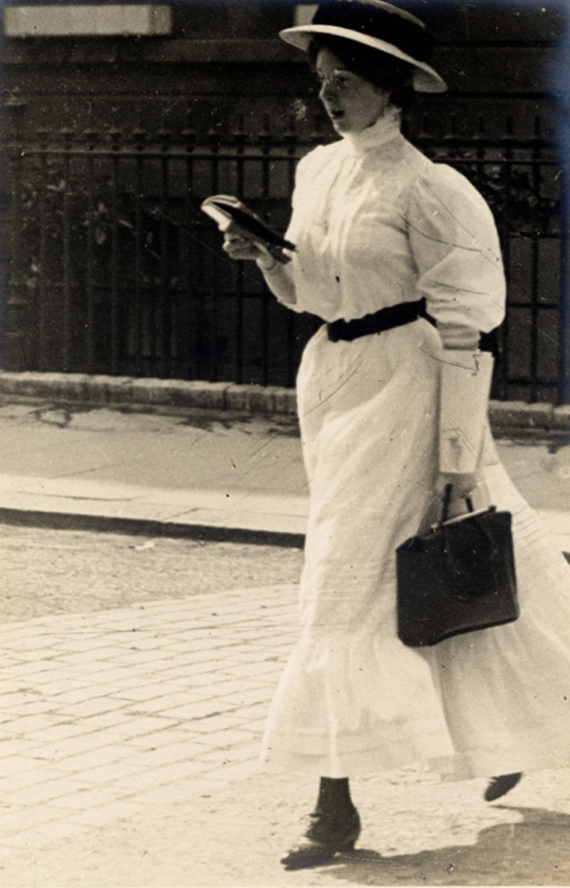A woman in a formal white dress with black handbag walks along the street in Kensington on June 15, 1908