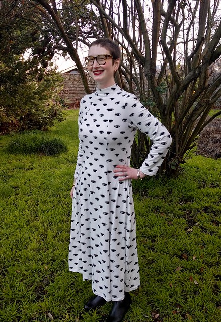 Woman stands in a garden. She wears a dramatic maxi dress in eye print.