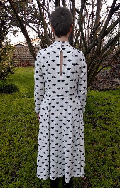 A woman stands in a garden. She is wearing a calf-length, full sleeve, high neck dress with black graphic eye print on white. Her back is to the camera.