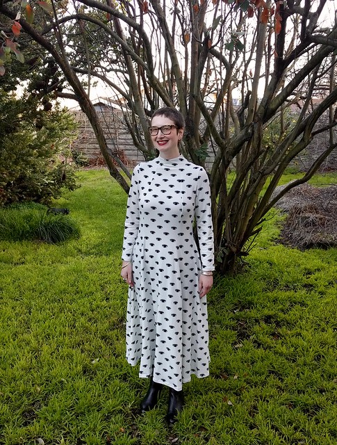 A woman stands in a garden. She is wearing a calf-length, full sleeve, high neck dress with black graphic eye print on white. She is smiling.