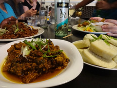 A black table covered with white plates of food.  In the foreground is a pile of dark brownish-red stew, sitting in a pool of liquid.  Next to this are sliced of peeled and boiled white yam, and behind is a plate of dark red rice and beans.  A wine bottle sits in the centre of the table.