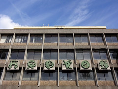 Six square plaques with light green mouldings on a grey background, attached to the side of a building with lots of blank windows.  They show stylised zodiac signs: a set of scales, a scorpion, a quiver of arrows, a goat, a face with flowing water, and two fish.