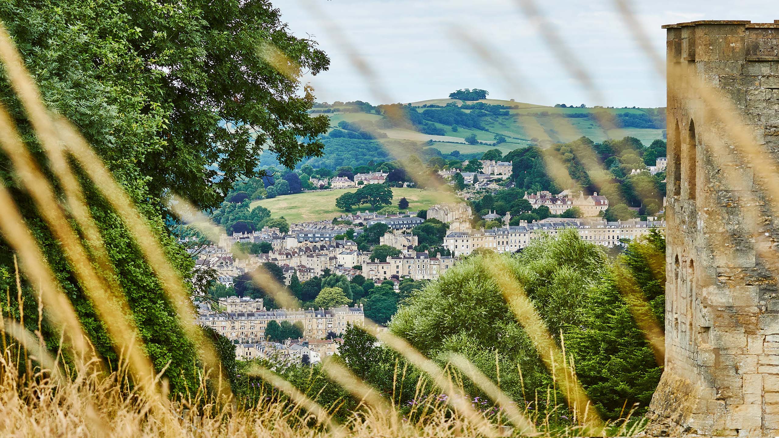 Peephole view of the city of Bath. The city, in the background, is framed by trees and shrubs on the left and Sham castle on the right. There are  rolling hills in the distant background and a very light-blue sky.