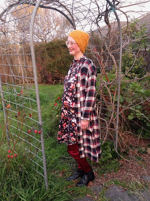 Woman stands in garden archway. She wears handknit yellow hat, checked flannelette long cardigan, black floral dress, burgundy velvet leggings and ankle boots.