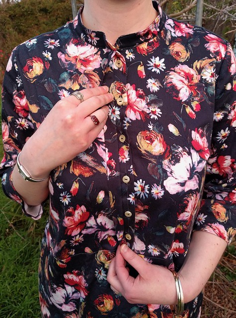 A close up of a woman's torso. She wears a black floral dress and holds open a concealed button band with two hands, revealing pearl-like buttons.