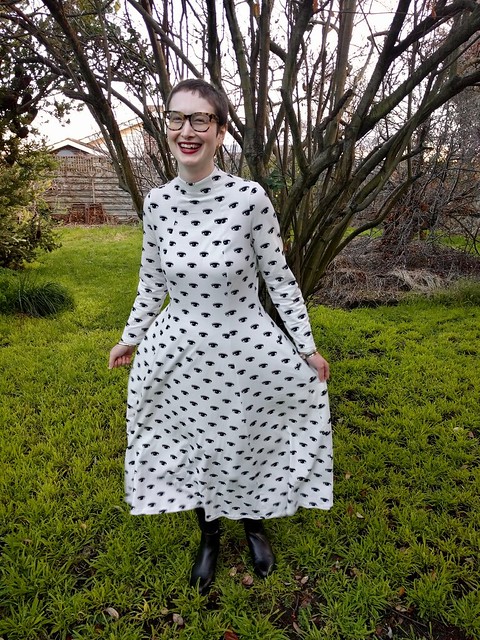 A woman stands in a garden. She is wearing a calf-length, full sleeve, high neck dress with black graphic eye print on white. She is smiling.
