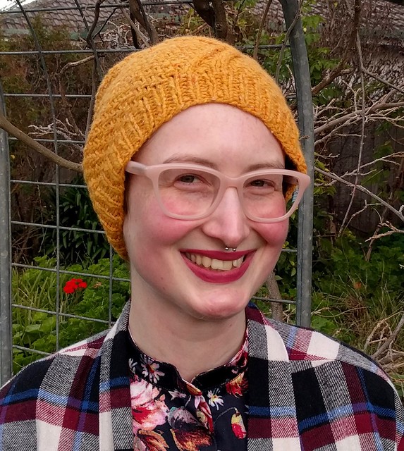 Woman stands in garden and wears a yellow handknit hat.
