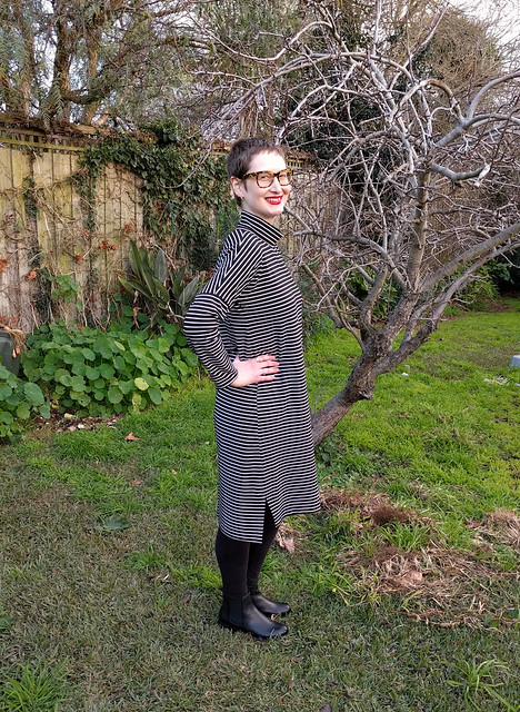 A woman stands in a garden. She wears a black and white striped dress in a cocoon shape with a turtleneck. She is smiling.
