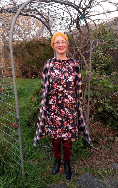 A photograph of a woman standing in a garden arch. She wears a long check cardigan, black floral dress, burgundy velvet leggings and a yellow hand knit hat. She is smiling.