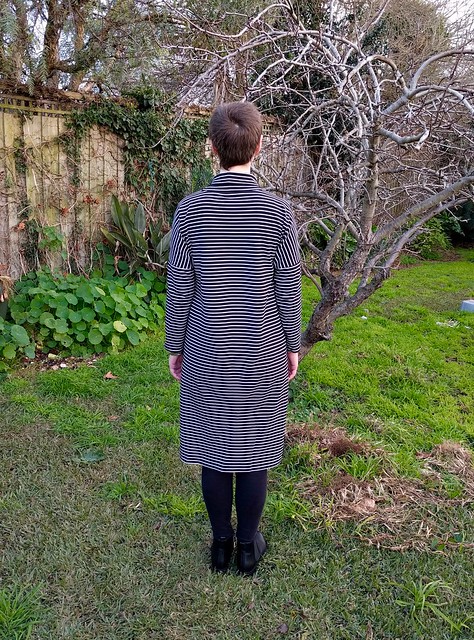 A woman stands in a garden. She wears a black and white striped dress in a cocoon shape with a turtleneck. Her back is to the camera.