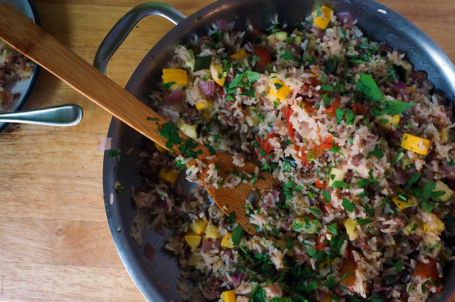 Overhead shot of a pan full of rice pilaf, the sunlight picking out bright shards of tomato and squash and the warm tone of the wooden spoon nestled in it.