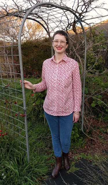 A woman stands in a garden archway. She wears a watermelon print button up shirt, jeans and ankle boots.
