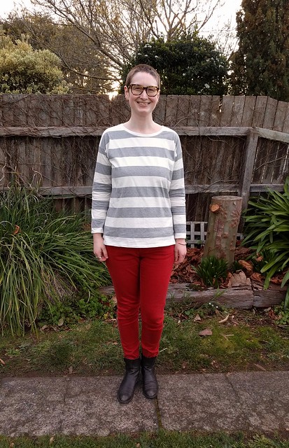 Woman stands against garden fence. She wears a long sleeve striped tee, red jeans and ankle boots.