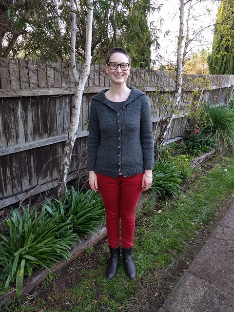 Woman stands against garden fence. She wears a grey handknit cardigan, red jeans and ankle boots.