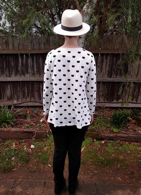 Woman stands against garden fence. She wears a long sleeve tee with cat head print, hat, black pants and ankle boots.