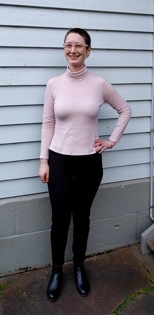 Woman stands in front of weatherboard house. She wears pink merino long sleeve tee, black pants and ankle boots.