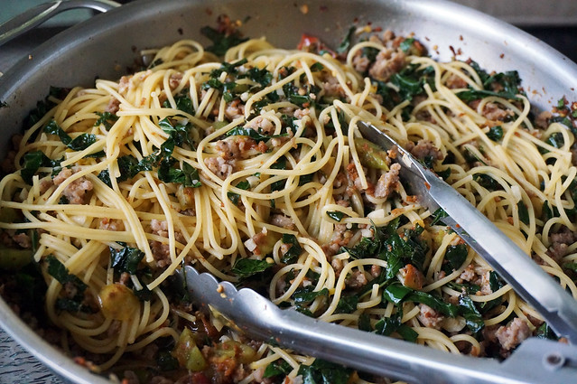 Closeup on spaghetti in a wide, deep pan, a pair of metal tongs ready to lift some out.