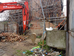 A red Kubota digger on a building site, poised as if just about to break the ground.  In the background is a partially demolished wall and a huge tangle of scaffolding.
