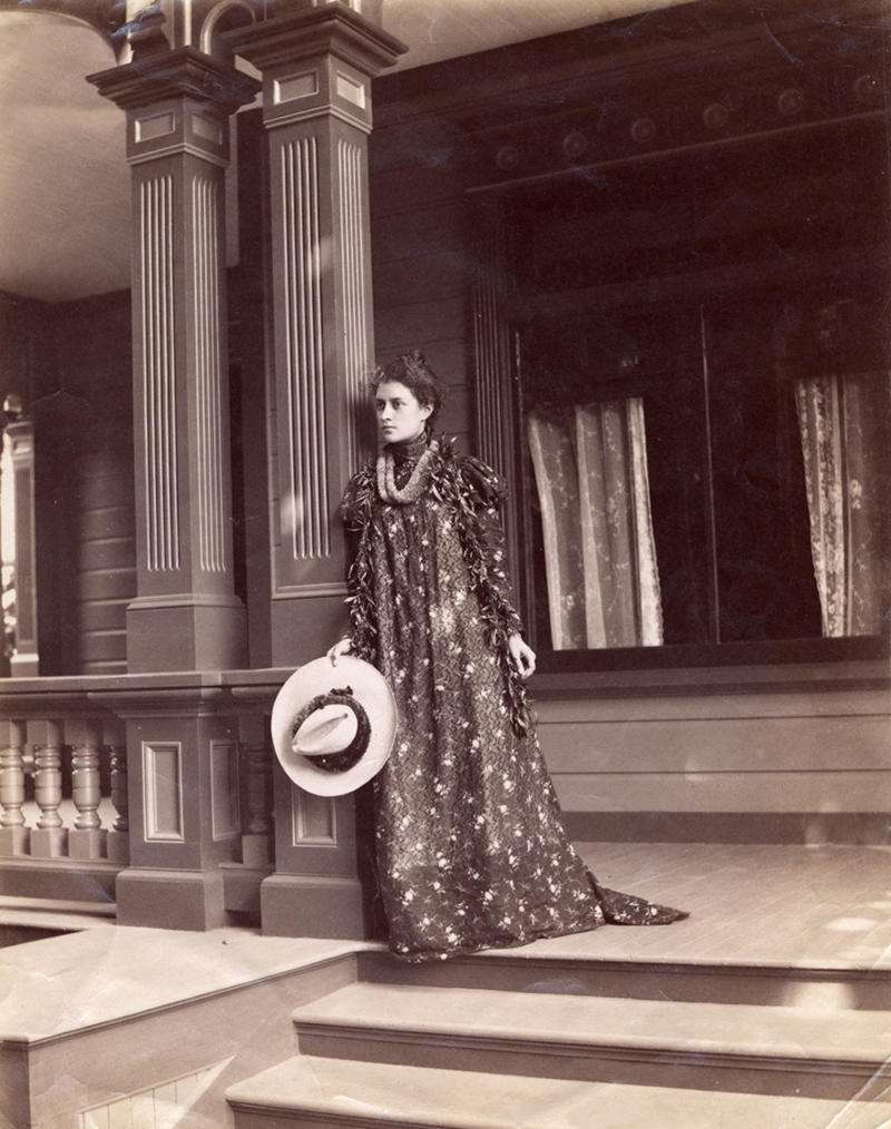 Princess Kaiulani standing on top of steps on the porch of her house at ʻĀinahau; wearing the holoku and a lei