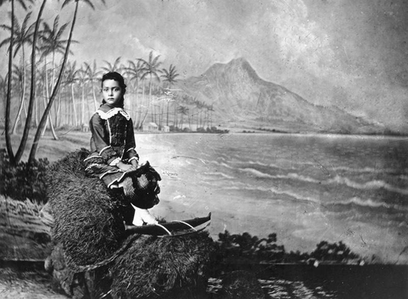 Kaiulani, approximately six years old seated holding hat with backdrop of Diamond Head & palm trees in a photo studio