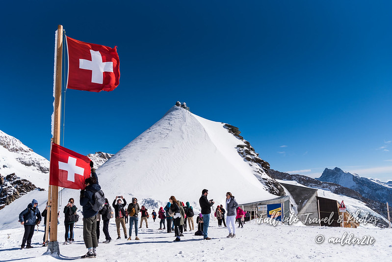 하얀 눈 밭 한 가운데에 빨간 스위스 깃발이 펄럭이고, 깃대 옆에서 사람들이 사진을 찍고 있다.