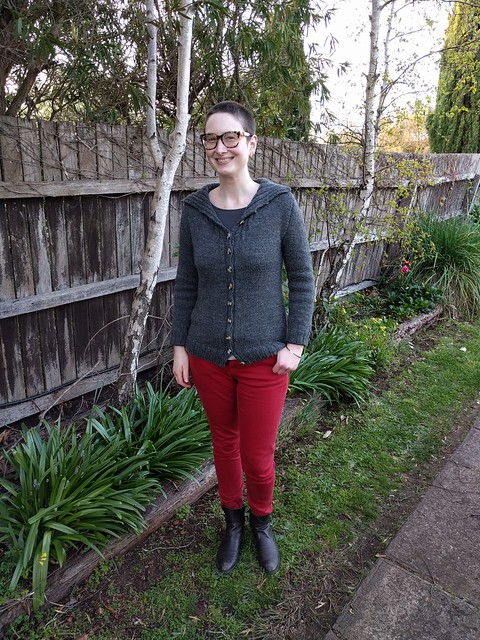 Woman stands against garden fence. She wears a grey handknit cardigan, red jeans and ankle boots.