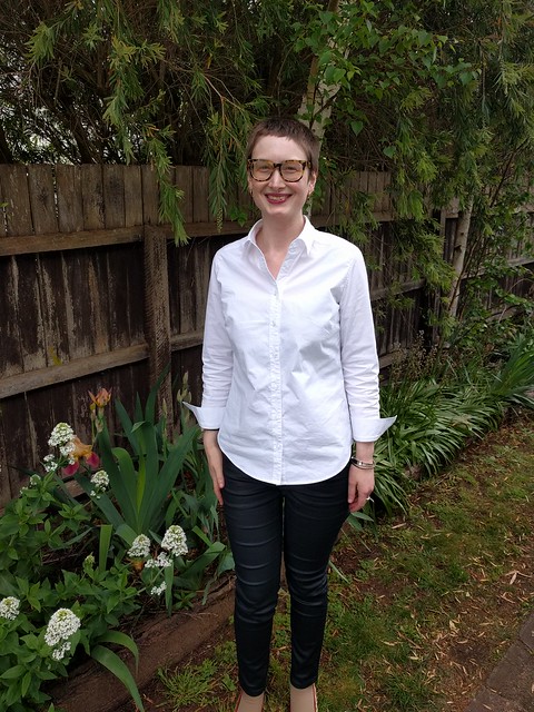 A woman stands against a garden fence. She wears a white button up shirt and black coated jeans.