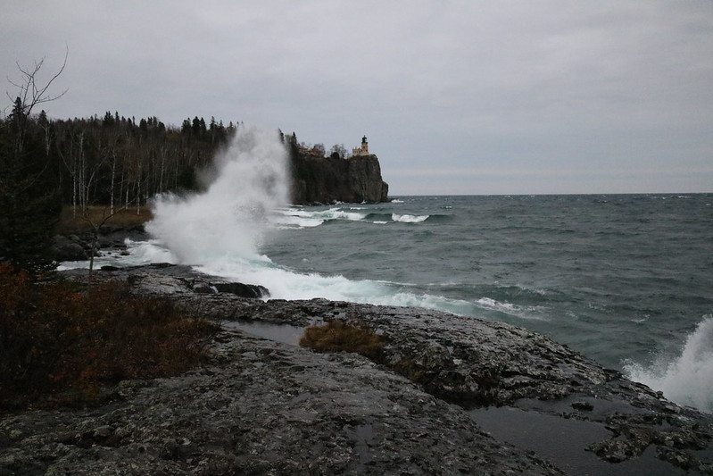 the lighthouse farther in the distance, a large flat rocky surface in the foreground, with a curved wave splashing to the left