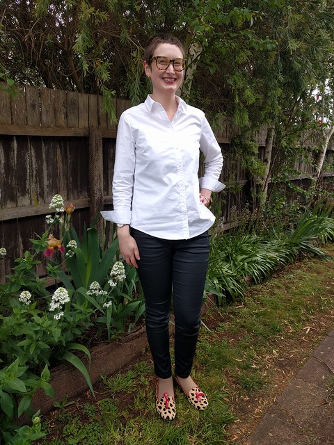 A woman stands against a garden fence. She wears a white button up shirt, black coated jeans and leopard print flats..