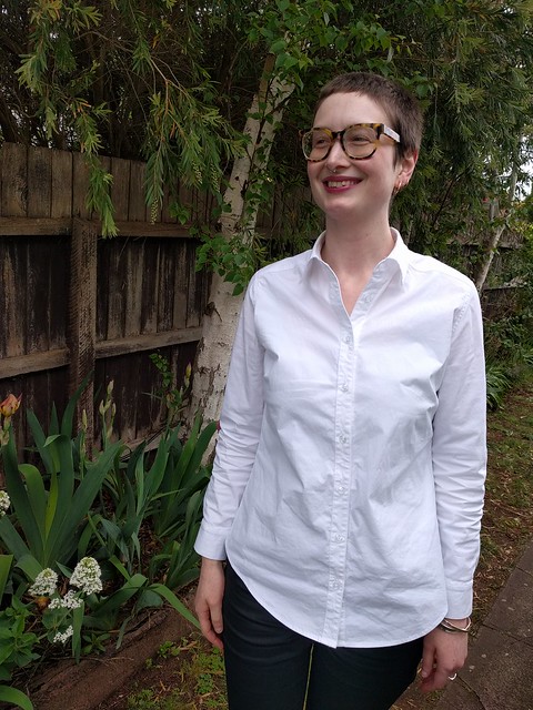 A woman stands against a garden fence. She wears a white button up shirt and black coated jeans.