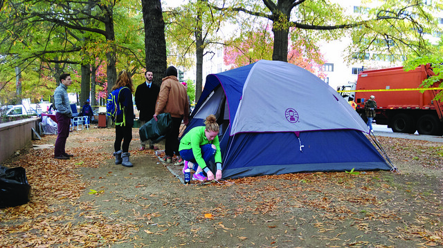 Photo of a young woman kneeling to pull up tent stakes while a group of people talk in the background. An orange trash truck is parked on the street in the background.