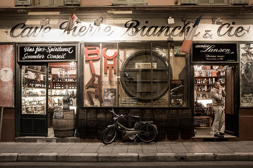 store boutique bottles evening late waiting france nice mobilette vintage smoking man flag signs pavement old wine champagne bike phoning color frontview 2wheels letters lights street sidewalk 1person candidphotography fulllength alpesmaritimes brown europe winery