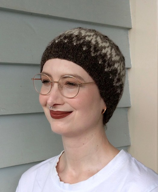 Woman's head in front of weatherboard house. She wears a handknit, colourwork hat in natural shades of brown and white.
