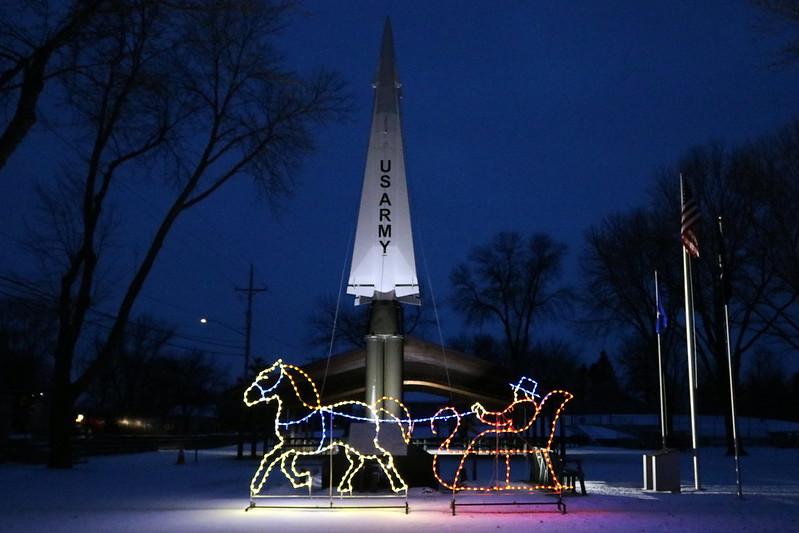 a wire horse pulling a man in a sleigh at the right, with a US Army statue in the background, at dusk