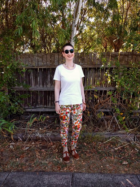 A woman stands in front of a garden fence. She wears sunnies, a white tee and tropical print slim fit pants.