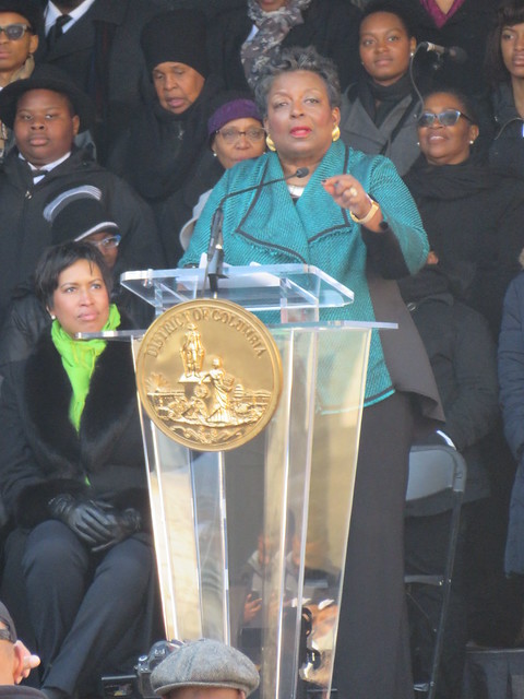 A photo of Cora Masters Barry speaking at a podium, with Mayor Muriel Bowser sitting beside her.