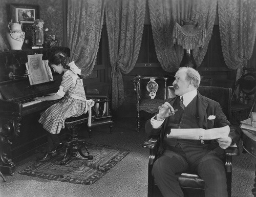 Antique-styled black-and-white composition of formally dressed older male teacher instructing young-adult female piano student