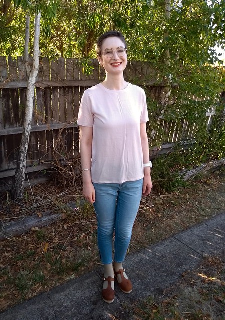 Woman stands in front of garden fence. She wears pink merino knit tee, light blue jeans and clogs.