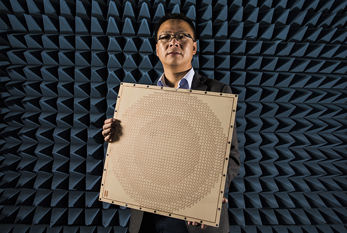 A man stands in front of a wall covered in foam spikes; he's holding a large square panel with both hands.