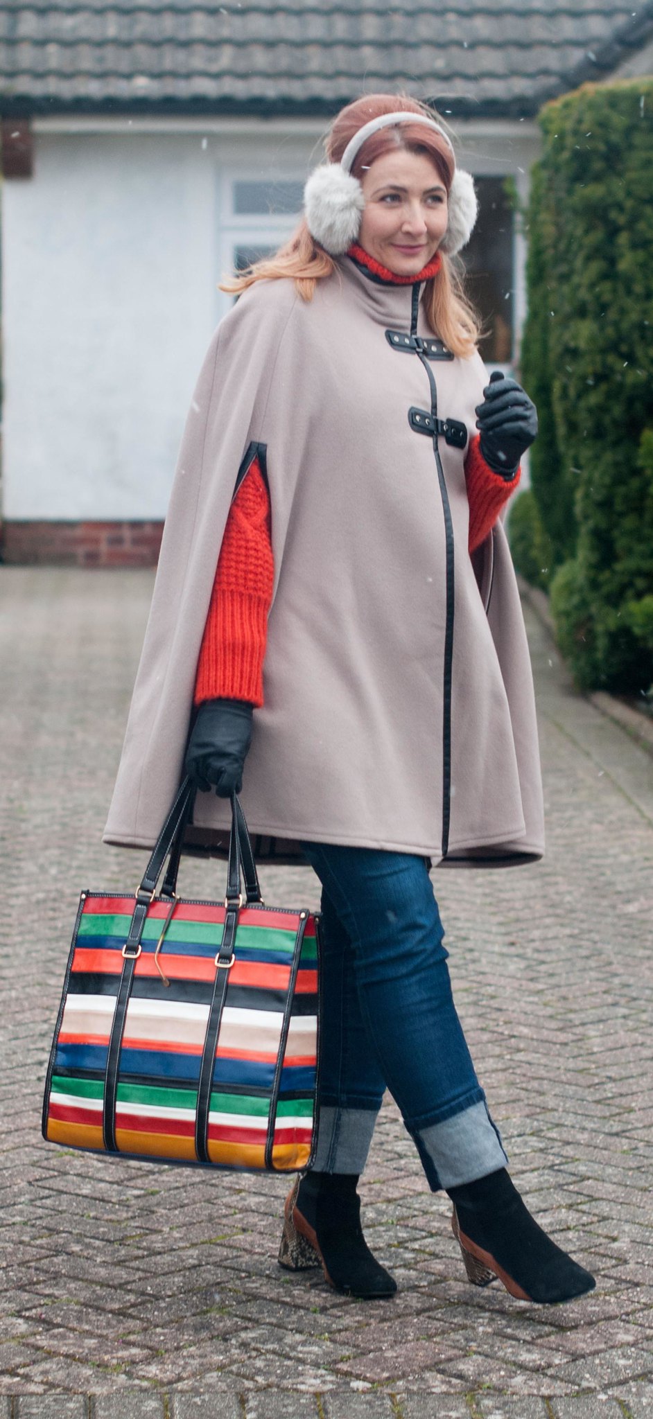 A Parisian Chic Winter Outfit, Over 40 Style \ camel wool cape \ ear muffs \ orange thick knit sweater \ rolled-hem jeans \ two-tone ankle boots \ rainbow-stripe tote bag | Not Dressed As Lamb