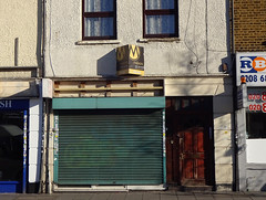 A ground-floor terraced shop with a green shutter covering the shopfront and an empty wooden frame above where the sign would have been.  A door to the right presumably leads to the upper floors.  An estate agent sign above reads “For Sale” and gives the name of the agent as “Malixons”.