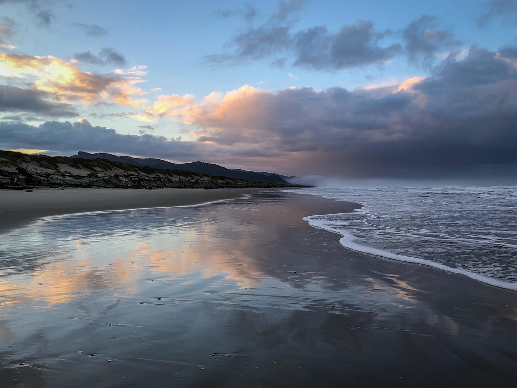 Kind of a seascape with a view from the self zone on the left to the coastal mountains on the left. Salmon tinged clouds reflected in wet beach sand.