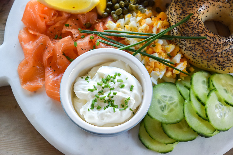 Whipped Feta in a white bowl on a board with salmon, capers, lemon, chopped egg, cucumber and bagels.